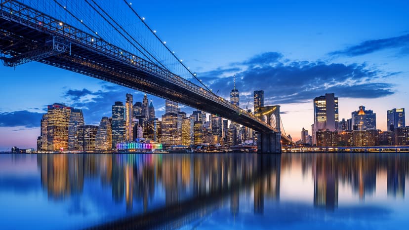 Brooklyn Bridge stretches over the East River at dusk with the illuminated Manhattan skyline and its reflection visible in the water.
