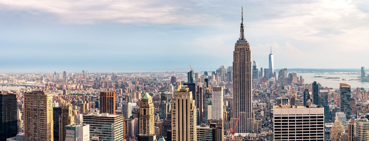 Panoramic view of the New York City skyline with the Empire State Building in the center and surrounding high-rise buildings under a partly cloudy sky.