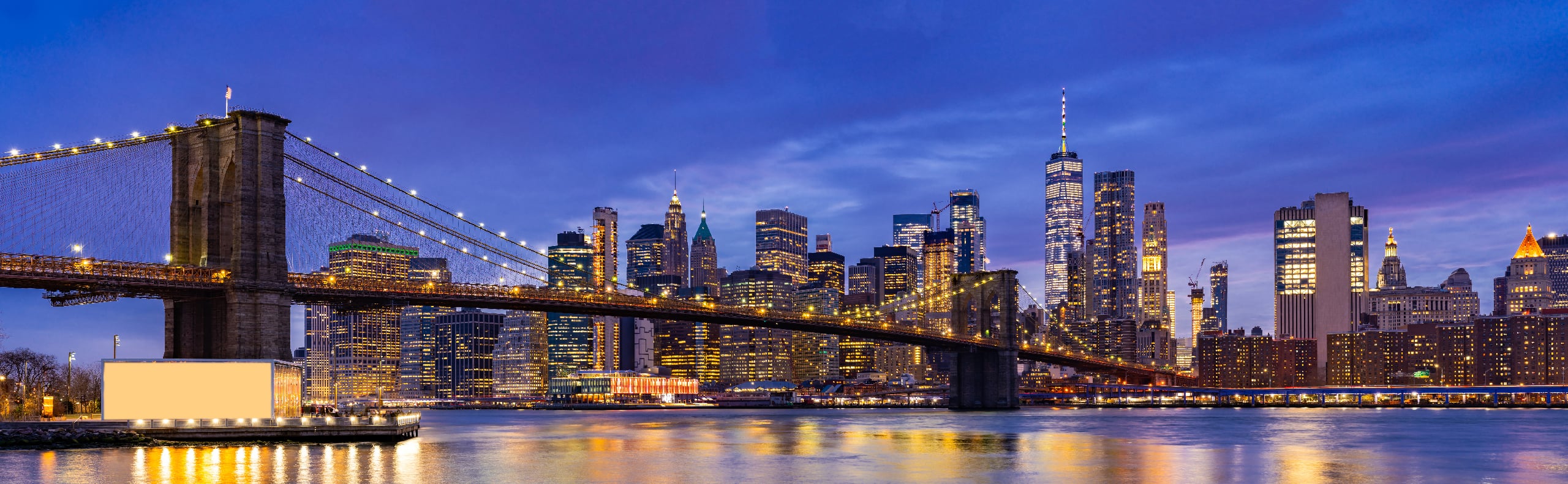 The Brooklyn Bridge spans over the East River with the illuminated Manhattan skyline in the background at dusk.